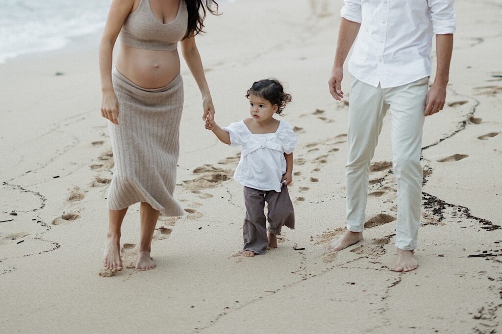 A young child walks along a beach holding the hand of her pregnant mother alongside her father, photo taken by a Hawaii maternity photographer