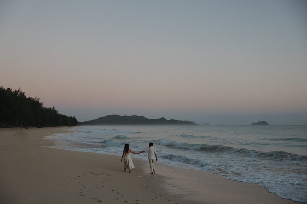 A photo taken by an Oahu engagement photographer of a couple walking hand in hand along Waimanalo Beach at sunrise