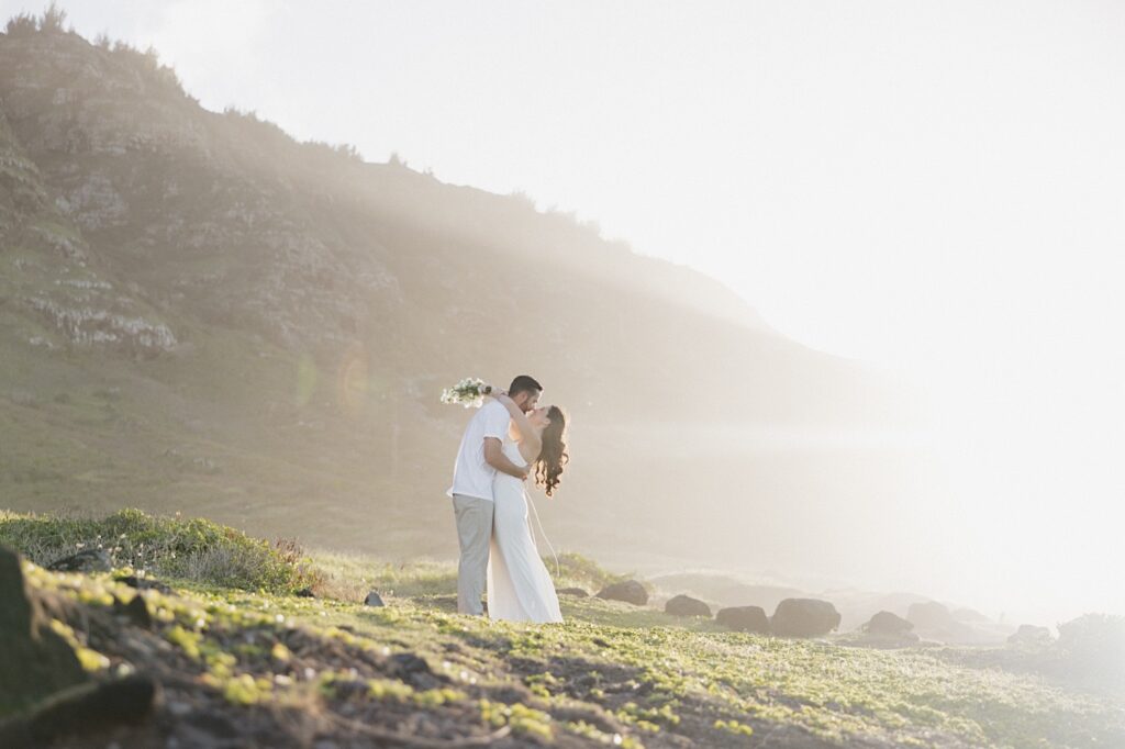 A bride and groom kiss on the side of a mountain in Hawaii for their elopement as the sun rises towards the right 