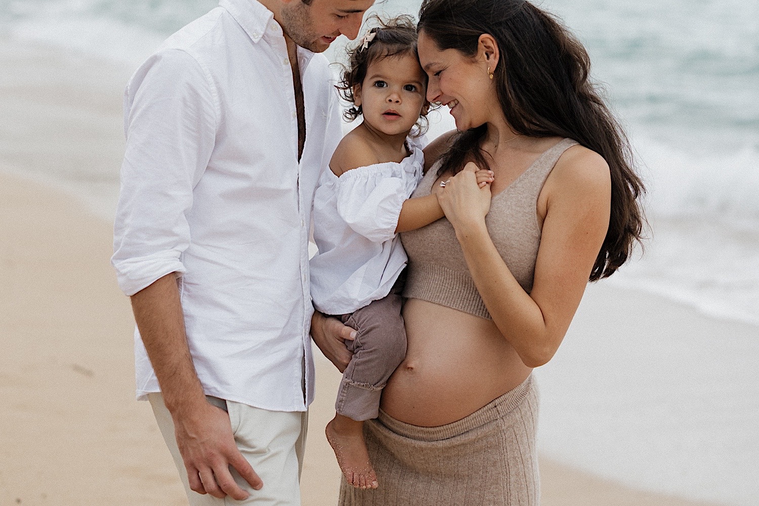 A child looks at the camera while being held between her pregnant mother and father on the beach, photo taken by a Hawaii maternity photographer