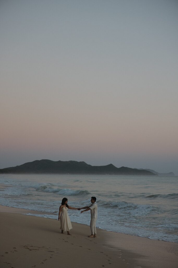 A couple dance together while holding hands on Waimanalo Beach on Oahu as the sun begins to rise, photo taken by an Oahu Engagement photographer