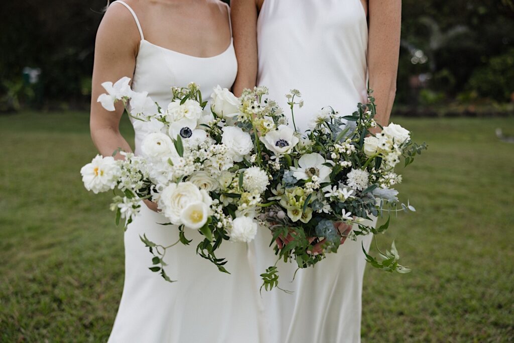 2 brides stand side by side and hold their bouquets towards the camera before their LGBTQ wedding day at Loulu Palm on Oahu