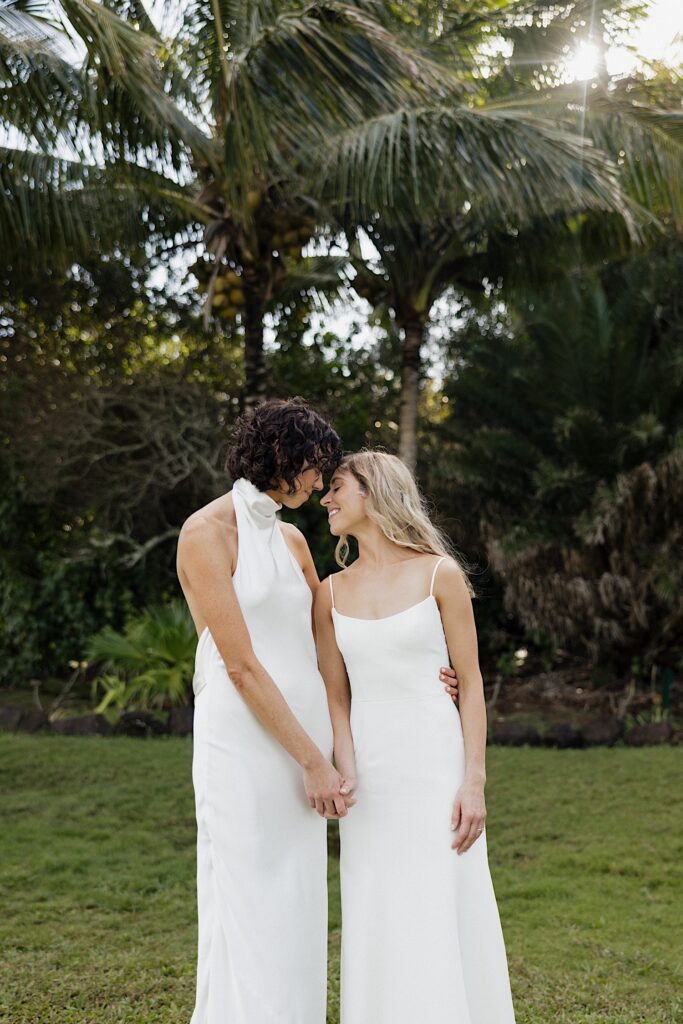 2 brides stand next to one another and hold hands while smiling and about to kiss in front of a palm tree at Loulu Palm on Oahu