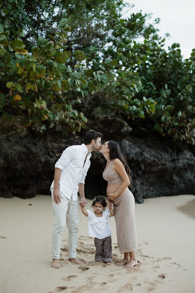A mother and father kiss one another while on a beach in Hawaii with their young daughter standing in between them holding their hands, the mother is holding her baby bump