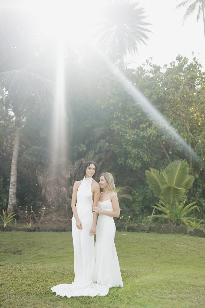 2 brides stand side by side in front of a palm tree under the sun before their wedding day at Loulu Palm on Oahu