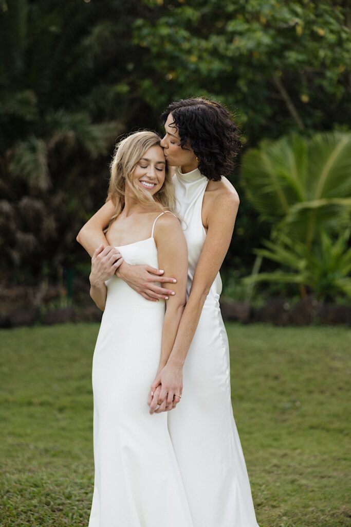 A bride smiles as the bride behind her hugs and kisses her head while at their wedding venue Loulu Palm on Oahu