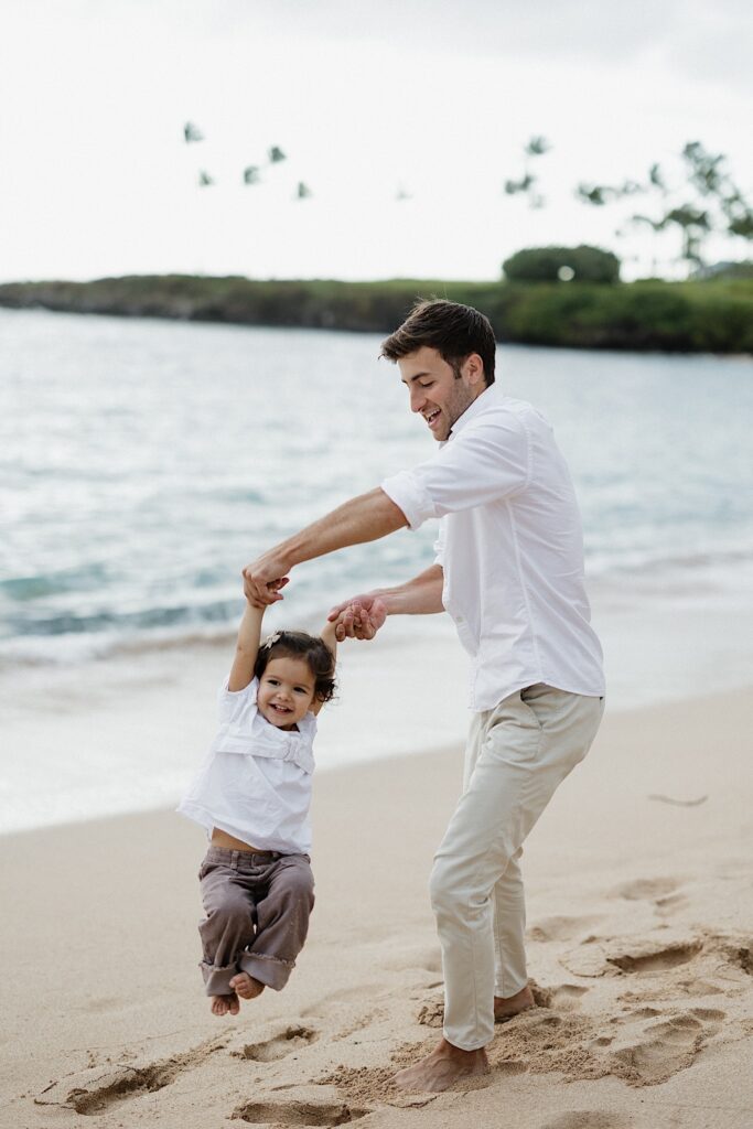 A young child smiles while being swung through the air by her father while the two play on a beach in Hawaii