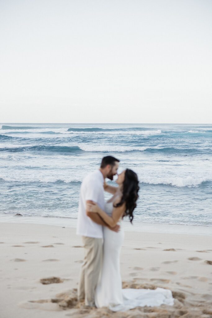 A bride and groom embrace on a beach in Hawaii and are about to kiss one another as waves crash behind them