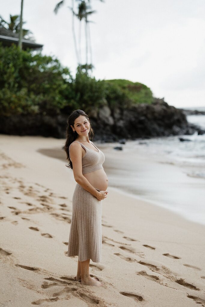 A pregnant woman stands on a beach in Hawaii during her maternity session and smiles at the camera while holding her baby bump