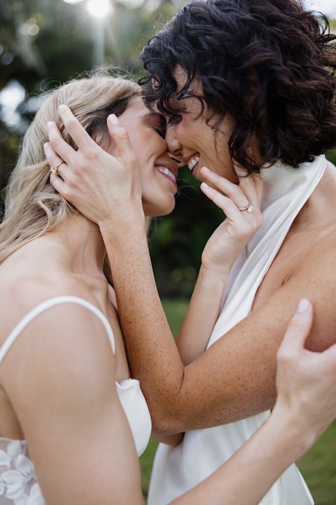 2 brides smile as they embrace and are about to kiss one another while the sun shines behind them