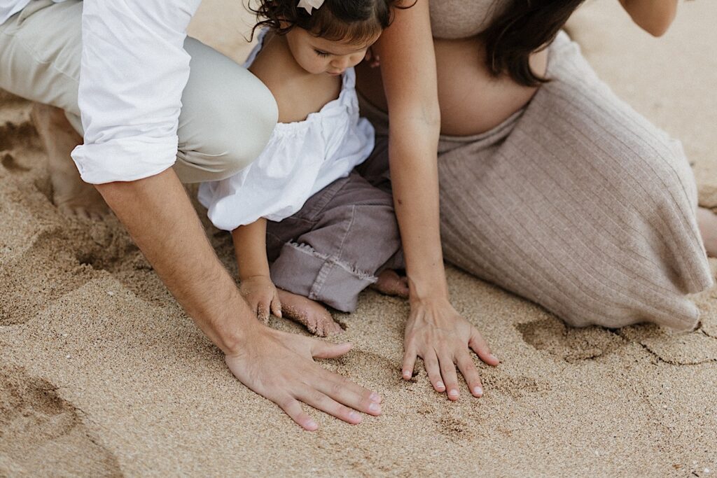 A child sits on a beach in Hawaii between her parents as they all put their hands in the sand, photo taken by a Hawaii maternity photographer