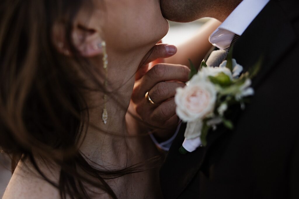 Close up photo of a wedding ring on a man's hand as he and the bride kiss one another and he touches her chin