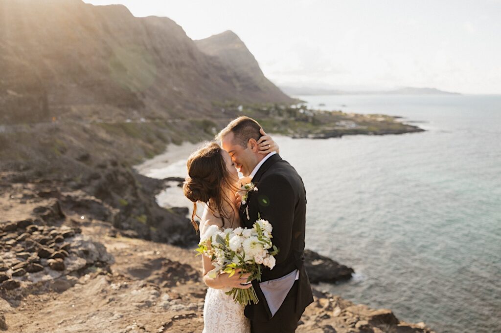 Atop Makapuu Lookout in Hawaii, a bride and groom kiss as the sun sets on them during their elopement