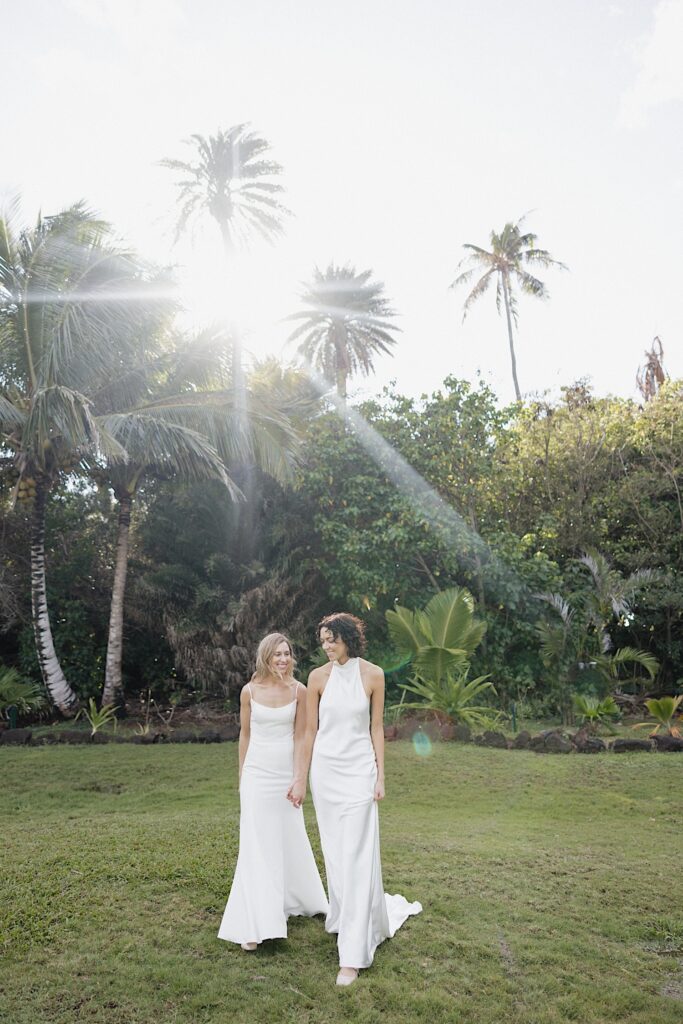2 brides smile while holding hands and walking towards the camera on the grounds of Loulu Palm on Oahu