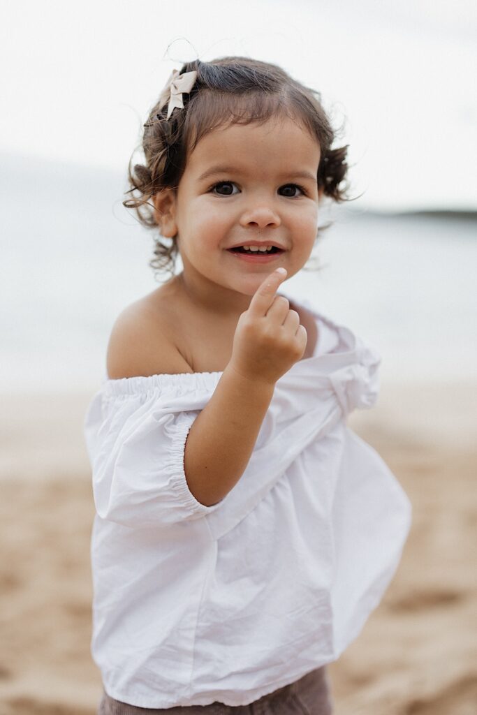A young child smiles at the camera while holding up one finger as she stands on a beach in Hawaii