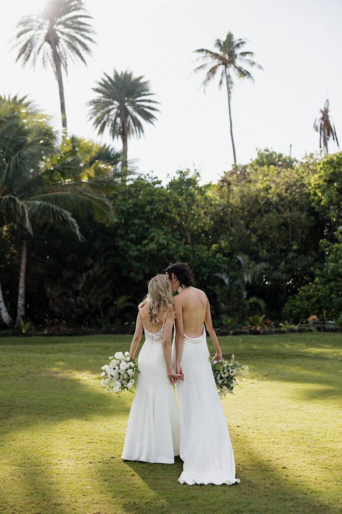 2 brides kiss while holding hands with their backs facing the camera, they stand in a clearing surrounded by palm trees at their wedding venue, Loulu Palm