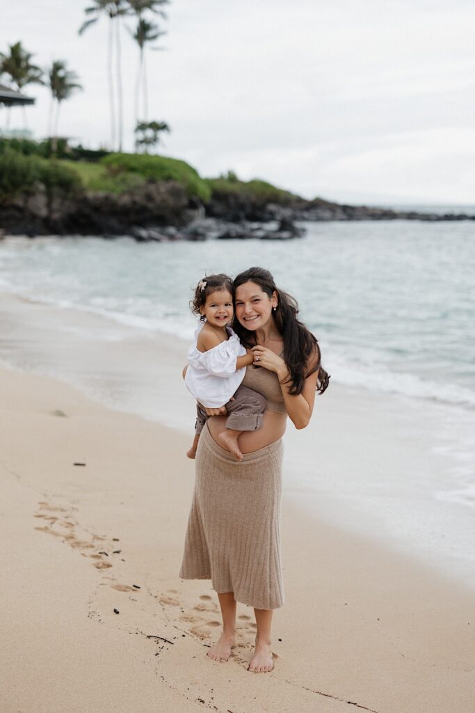 A pregnant mother smiles at the camera while holding her daughter in her arms as they stand on a beach in Hawaii