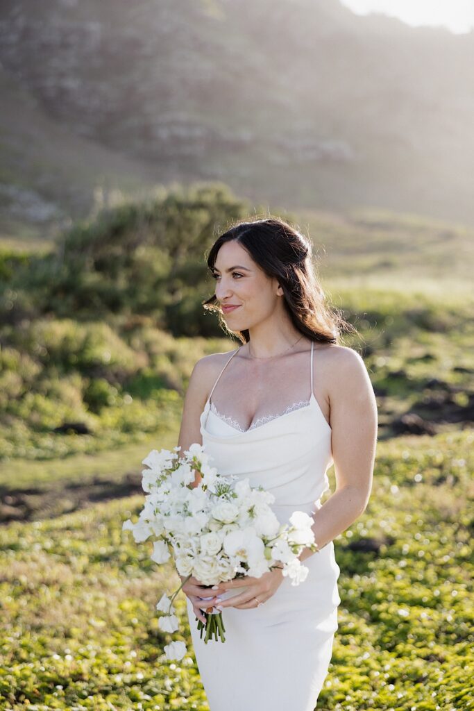 A bride holding her flower bouquet stands in a clearing in front of a mountain as the sun rises behind her