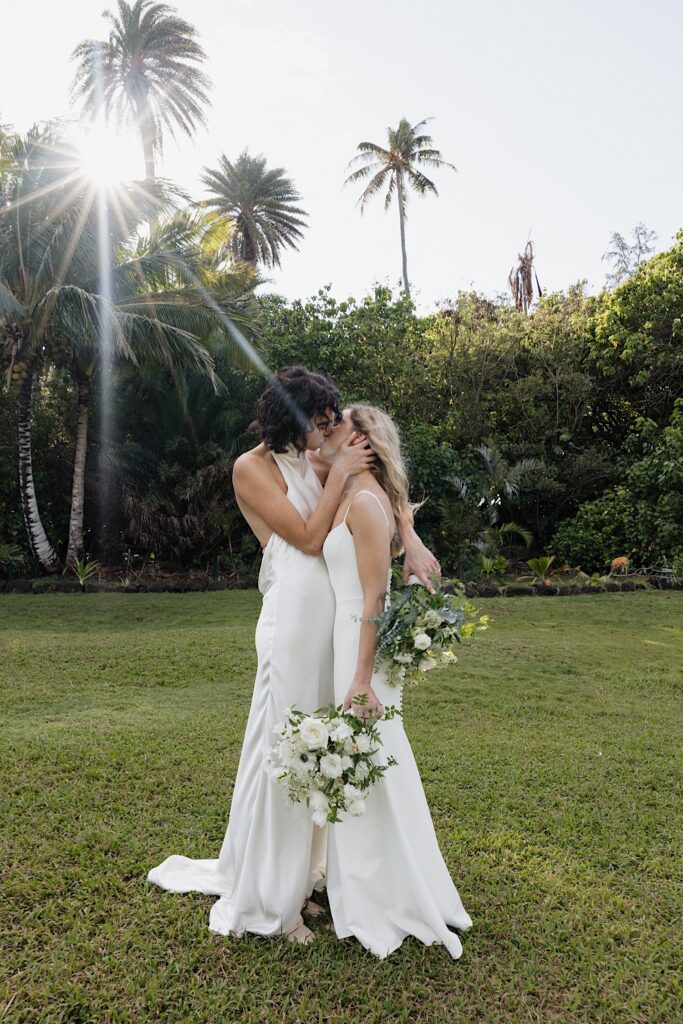 2 brides kiss one another while standing in a grass clearing under the sun at their wedding venue, Loulu Palm