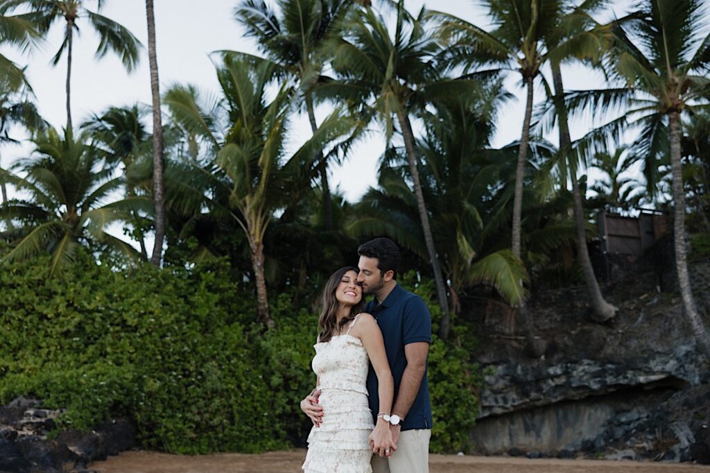 A pregnant woman smiles while being kissed on the cheek from behind by a man while on a beach in Hawaii, photo taken by a Hawaii maternity photographer