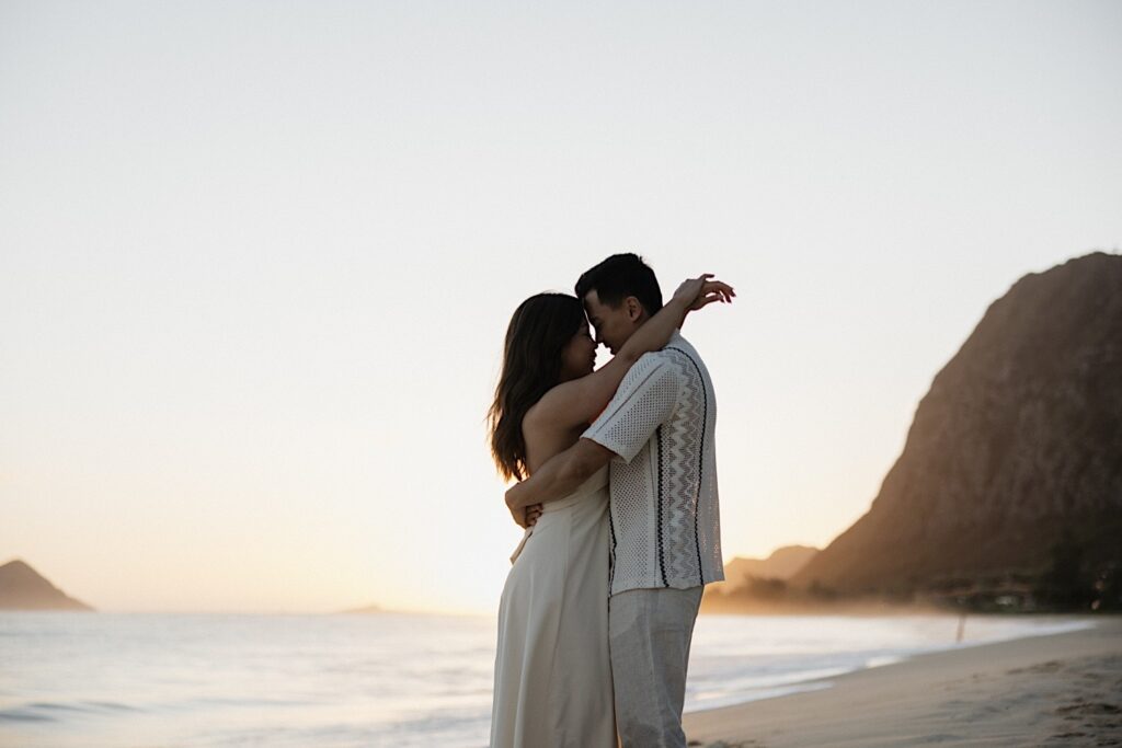A couple embrace one another while the sun rises behind them on Waimanalo Beach, photo taken by an Oahu engagement photographer