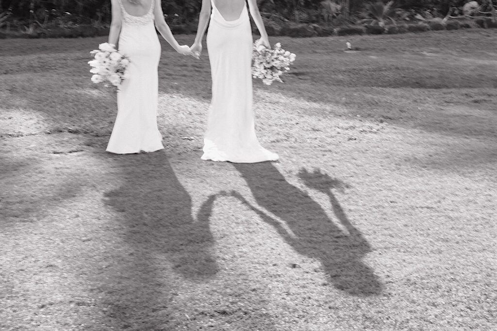 Black and white photo of 2 brides holding hands and facing away from the camera in a grass clearing before their LGBTQ wedding day at Loulu Palm on Oahu