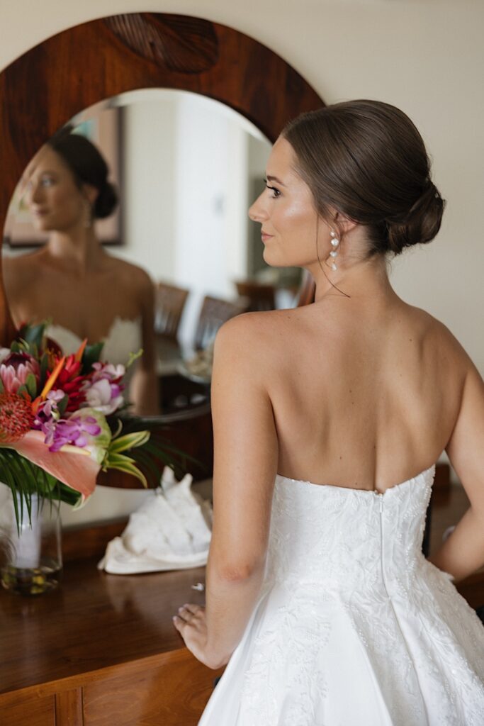 A bride stands in a mirror and looks over her shoulder after getting ready for her wedding day