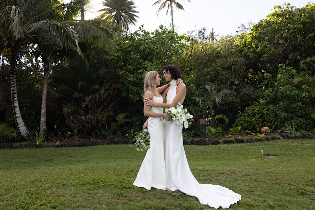 2 brides embrace and smile at one another while standing in a grass clearing at their wedding venue, Loulu Palm on Oahu prior to their LGBTQ wedding