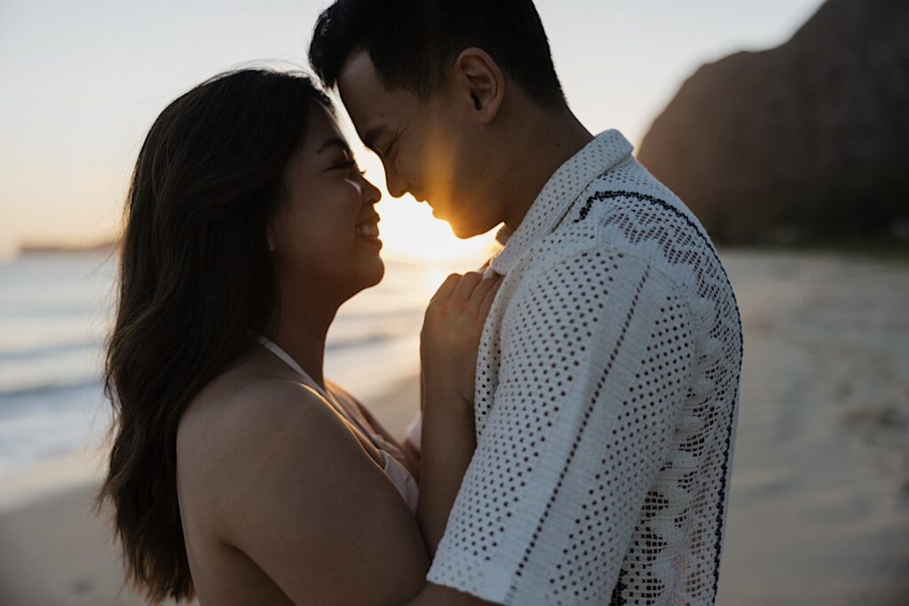 A couple hug and smile at one another while on Waimanalo Beach as the sun rises in the background between them, photo taken by an Oahu engagement photographer