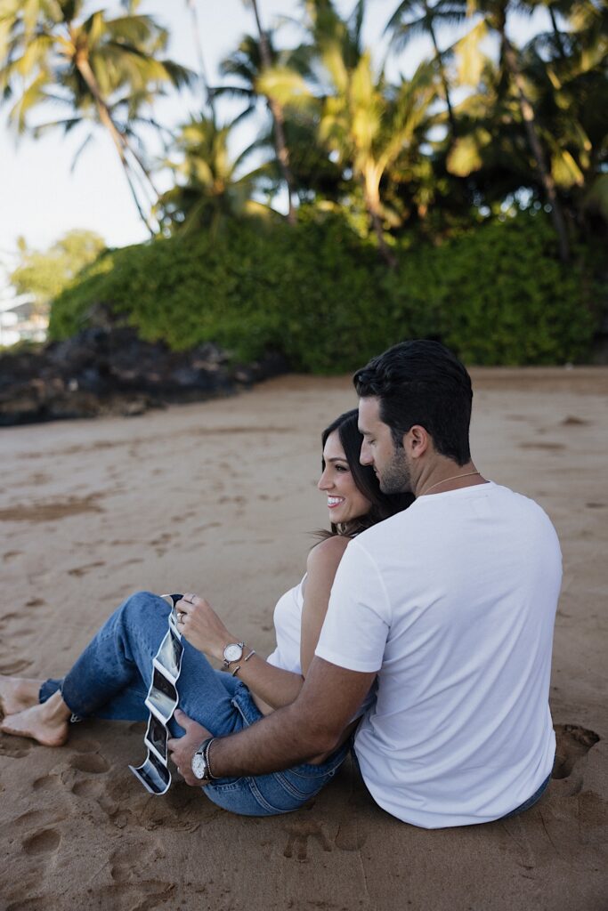 A couple sit together on a beach in Hawaii and smile while the woman holds her ultrasound photos in one hand