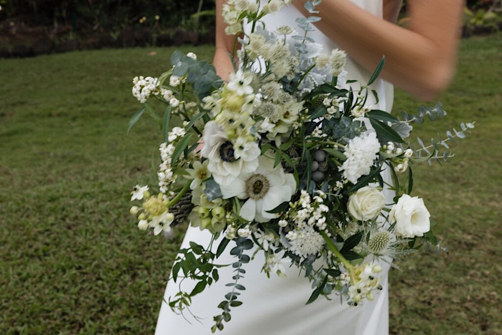 A bride holds her white floral bouquet towards the camera while standing in a grass clearing at her wedding venue, Loulu Palm