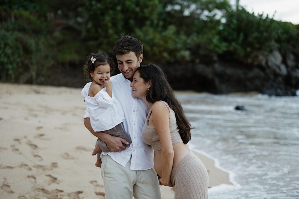 A young child smiles at the camera while being held by her father as the pregnant mother smiles next to them while the three are on a beach in Hawaii, photo taken by a Hawaii maternity photographer