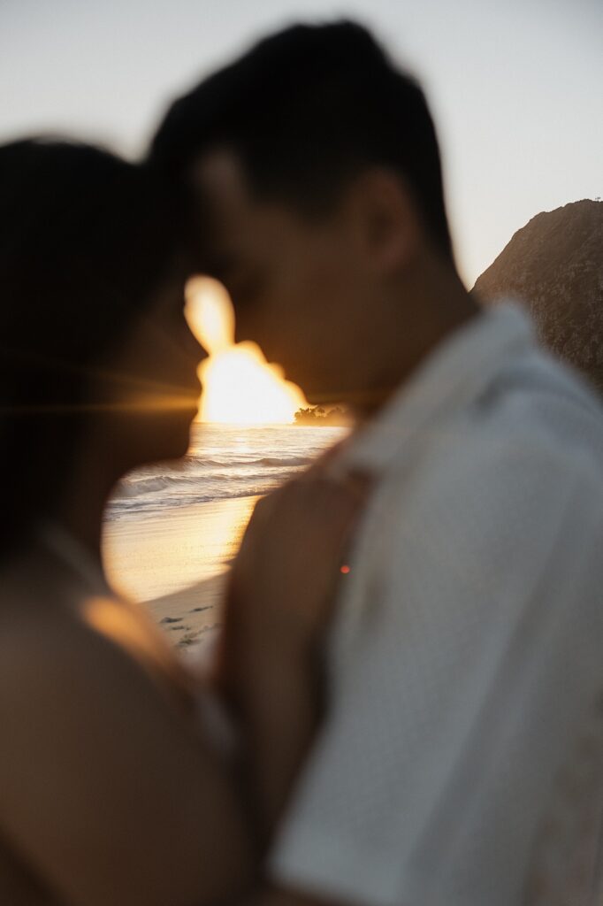 A couple embrace one another and touch foreheads together while out of focus, in between them is the ocean at sunrise in focus, photo taken by an Oahu Engagement photographer