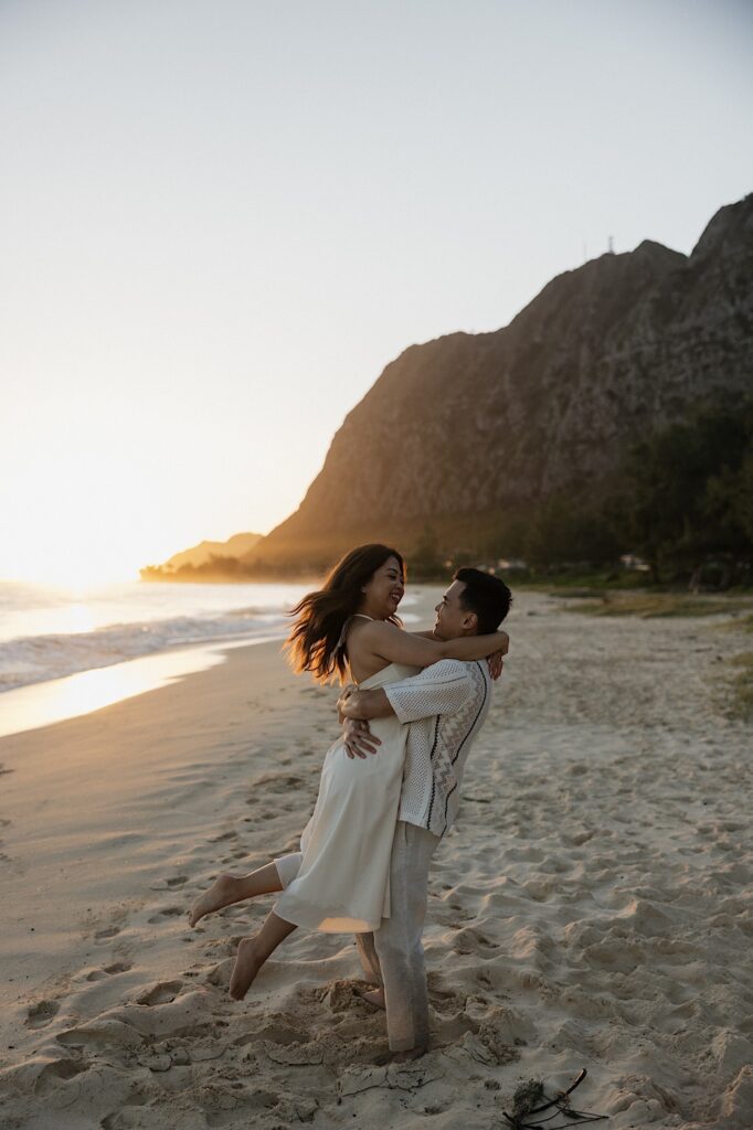 A man hugs a woman as he lifts her in the air while they both smile standing on Waimanalo Beach at sunrise, photo taken by an Oahu Engagement photographer