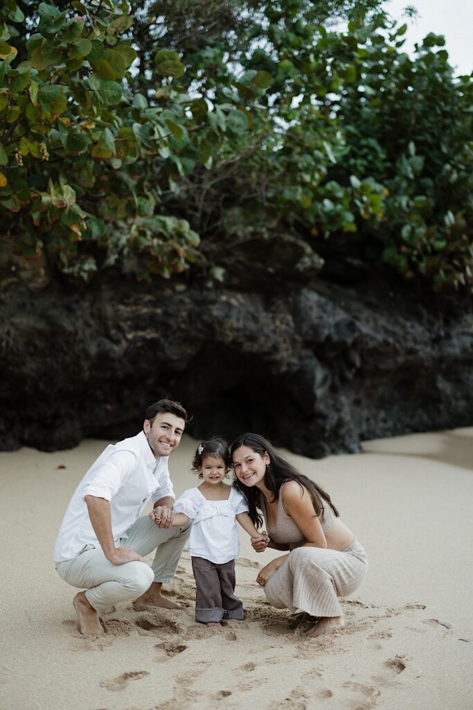 A young child stands on a beach in Hawaii between her two parents as the three of them smile at the camera