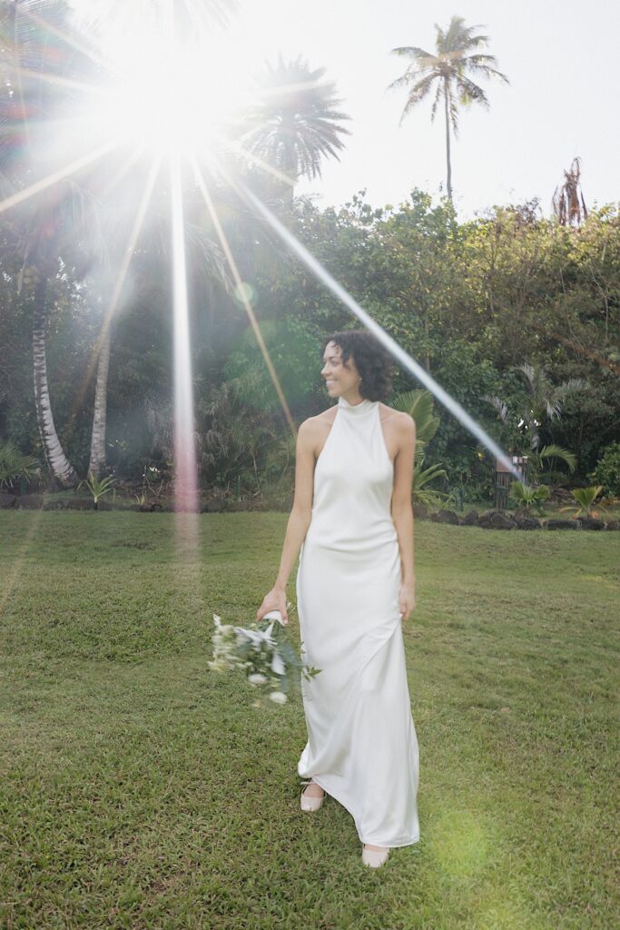 A bride smiles while holding her bouquet in a grass clearing as the sun shines behind her