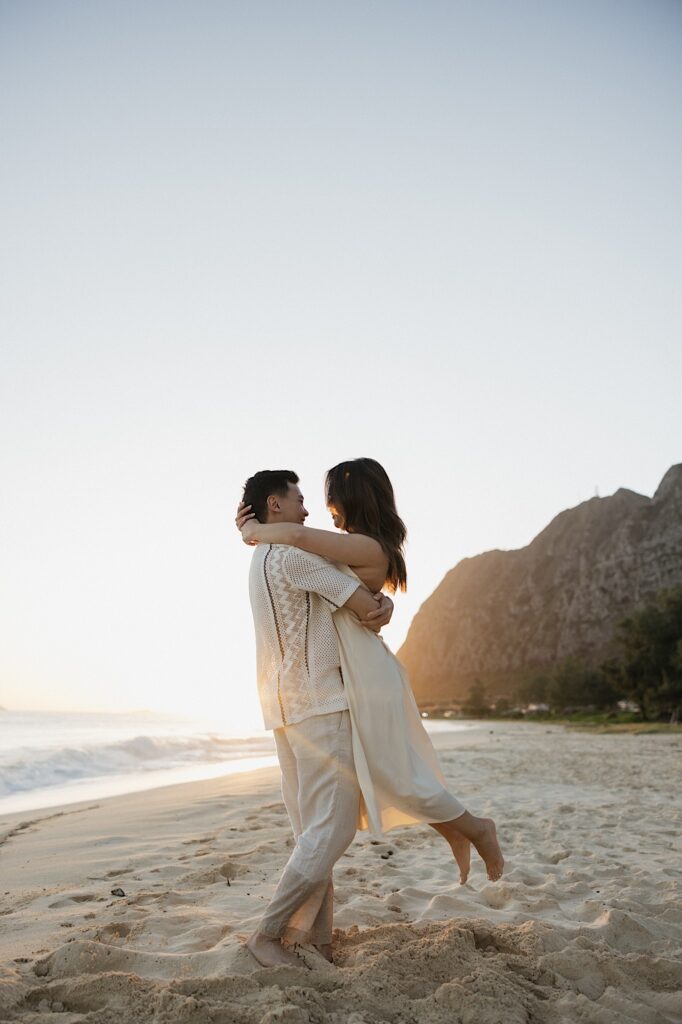 A man holds a woman in the air while they smile at each other during sunrise at Waimanalo Beach, photo taken by an Oahu Engagement photographer