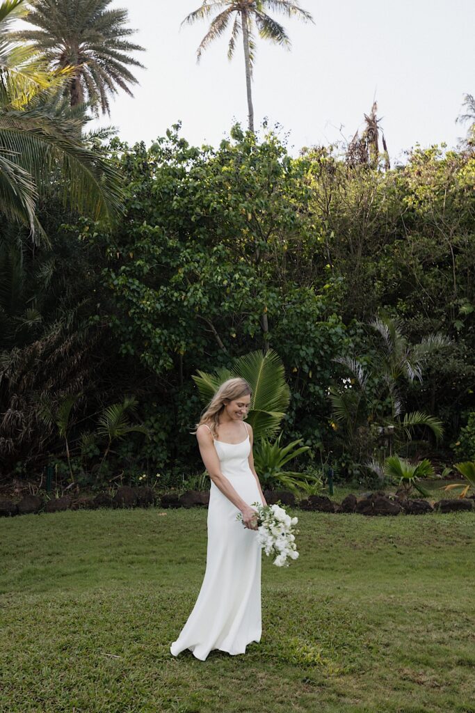 A bride smiles while looking down at her flower bouquet while standing in a grass clearing at her wedding venue, Loulu Palm