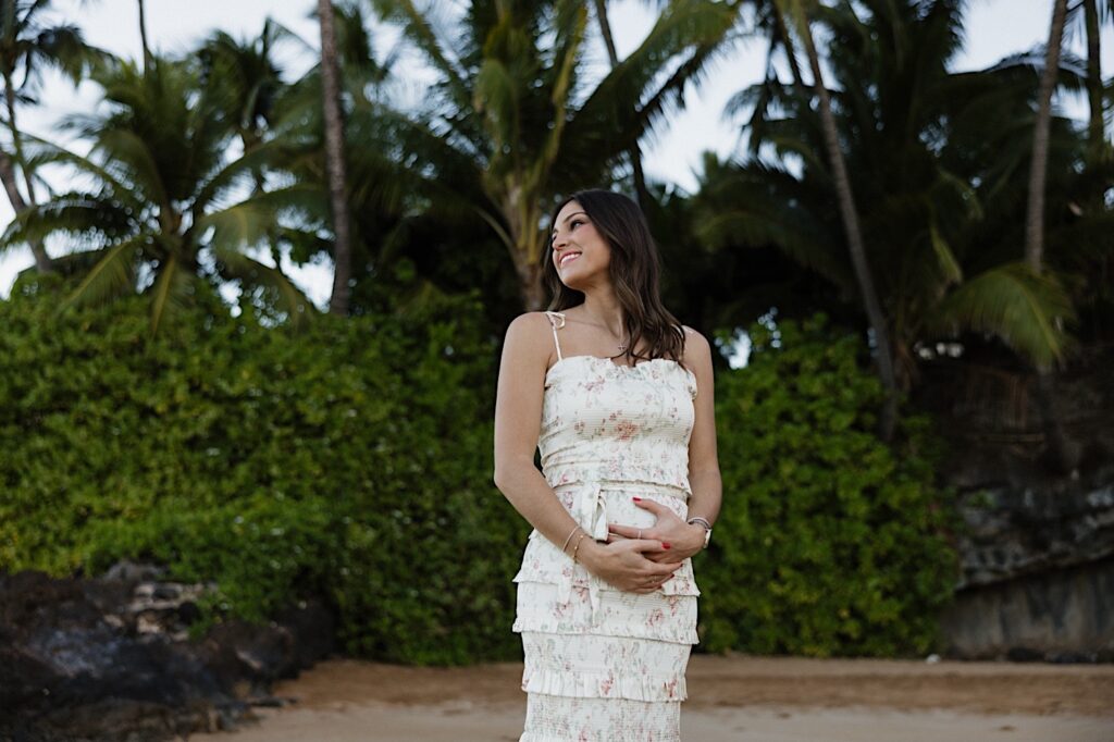 A woman smiles while holding her stomach during her maternity session on a beach in Hawaii, photo taken by a Hawaii maternity photographer
