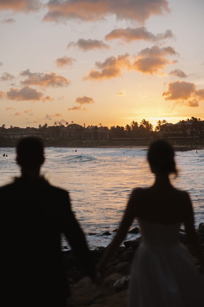 A bride and groom hold hands and walk towards the ocean while in Hawaii as the sun sets in front of them
