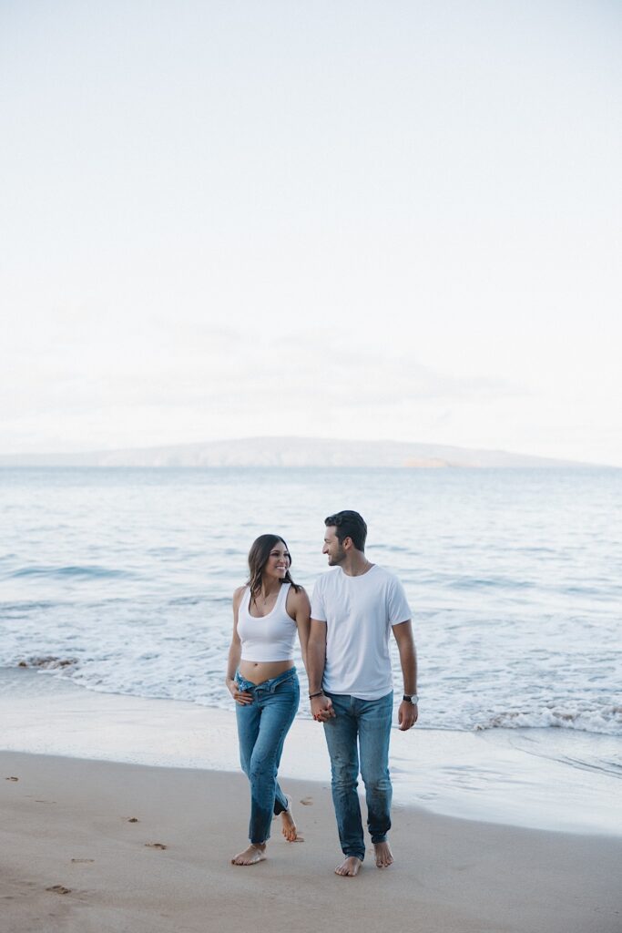 A couple walk hand in hand on a beach in Hawaii away from the ocean while smiling at one another