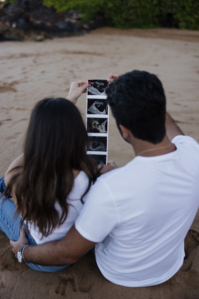 A couple sit together on a beach in Hawaii facing away from the camera as they look at ultrasound photos together
