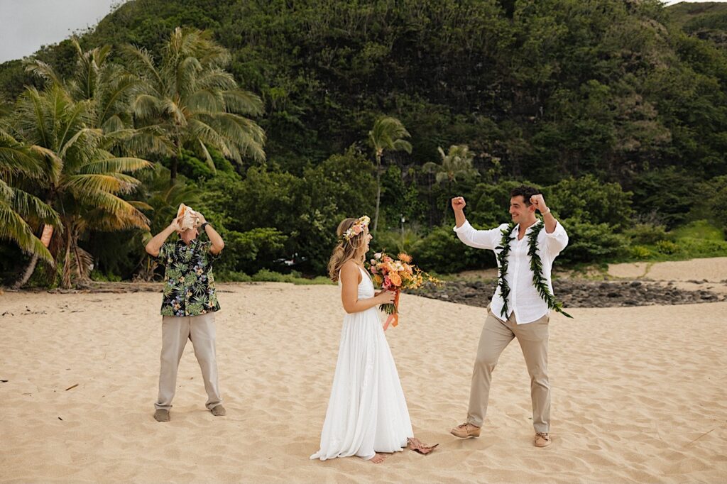A groom raises his firsts in the air as the bride smiles while their officiant blows into a shell to celebrate their elopement  on a beach in Hawaii