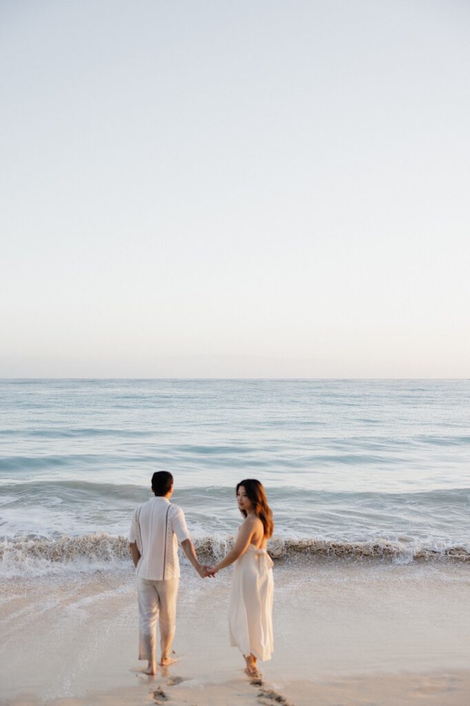 A woman looks back to the camera while holding hands with a man as they walk into the ocean together at sunrise