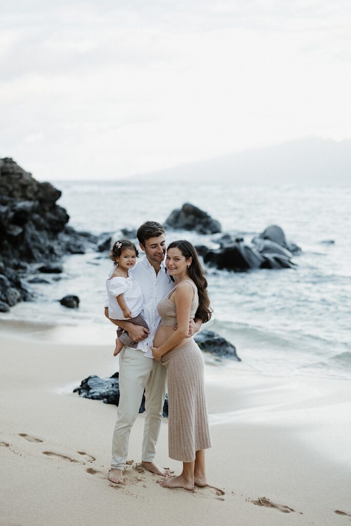 A pregnant mother smiles at the camera while her husband smiles at her and holds her and their young daughter while on a beach in Hawaii
