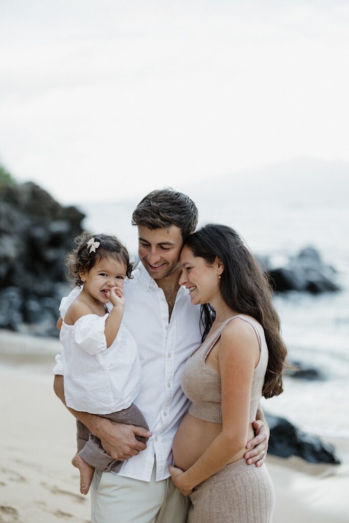 A young girl smiles at the camera while being held by her father who is standing next to the smiling mother on a beach in Hawaii