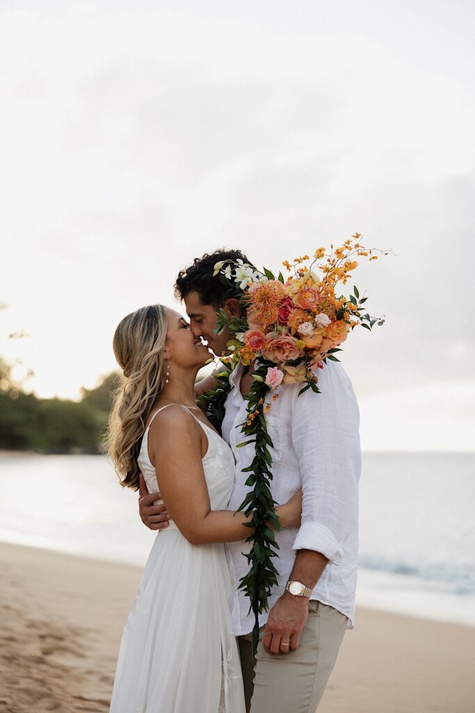 A bride and groom on a beach in Hawaii embrace and lean in to kiss one another at sunset