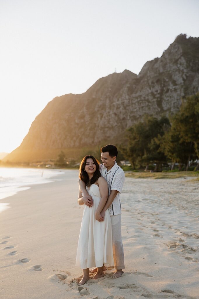 A couple smiles while the man hugs the woman from behind while on Waimanalo Beach at sunrise, photo taken by an Oahu Engagement photographer