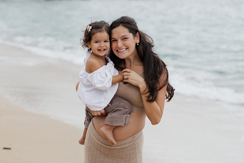 A pregnant mother smiles at the camera while holding her smiling daughter as she stands on a beach in Hawaii, photo taken by a Hawaii maternity photographer