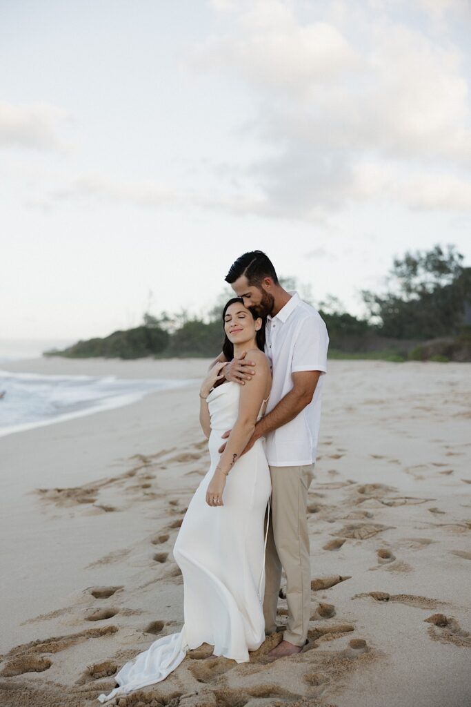 A bride smiles as the groom hugs her from behind while the two stand on a beach in Hawaii